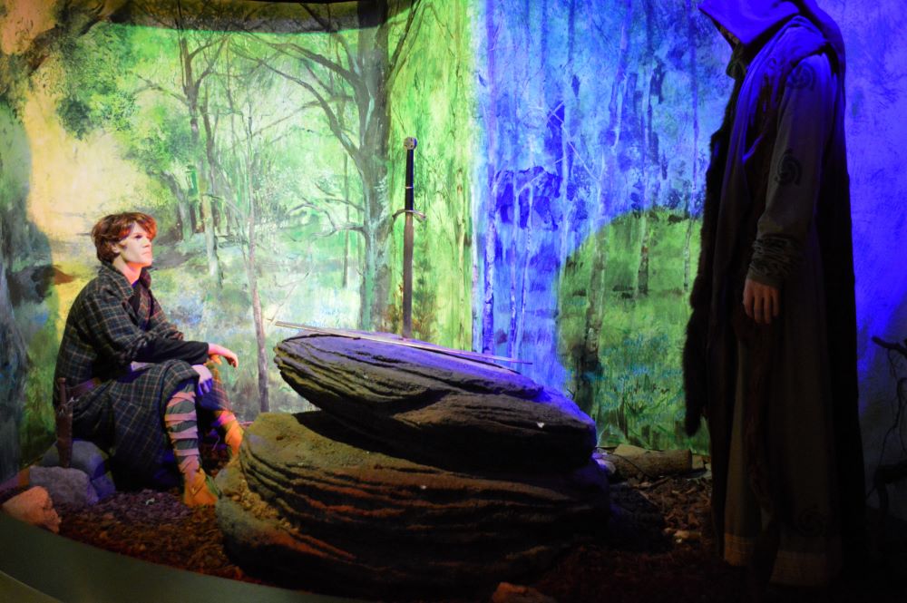 un mannequin représentant un jeune homme fait face à un mannequin représentant un mage, avec entre eux une épée plantée dans un rocher