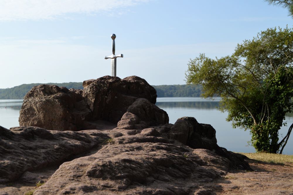 une épée plantée dans un rocher au premier plan, avec un lac en arrière plan