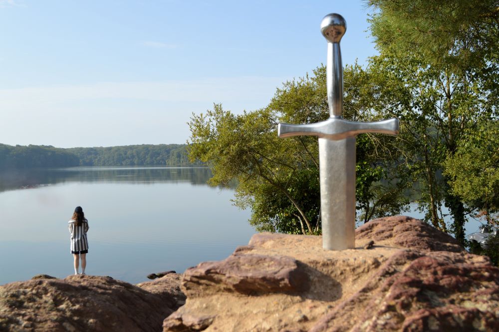 une épée plantée dans un rocher au premier plan, avec un lac en arrière plan