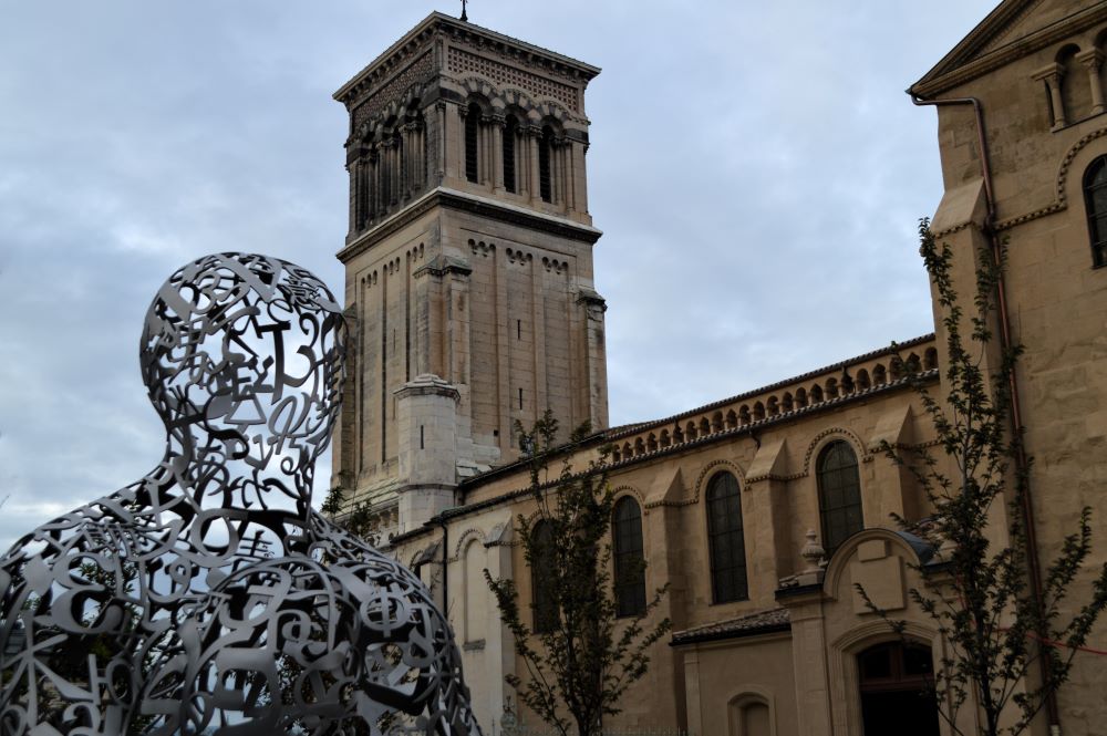 Sculpture en lettres d'acier assemblées pour former une silhouette humaine devant une cathédrale 