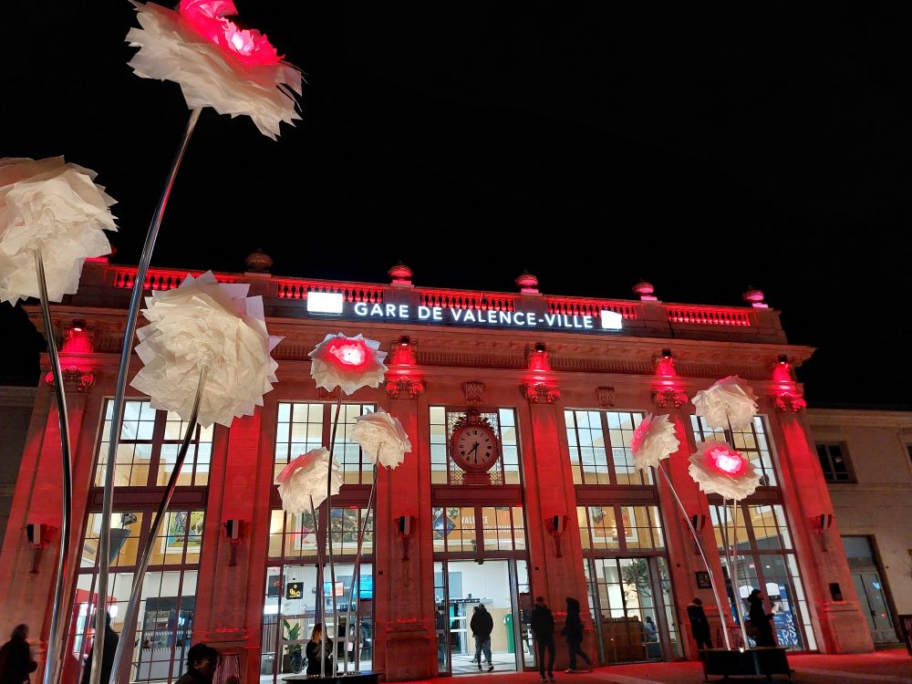 de nuit, des fleurs géantes en papier sont installées sur le parvis d'une gare dont la façade est éclairée en rouge
