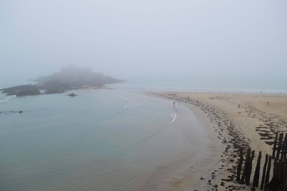 une plage dans le brouillard à Saint Malo où on devine les rochers et bâtiments du Fort National