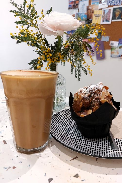 un grand verre de latte et un muffin sur une table décorée d'un petit bouquet de mimosa et renoncule