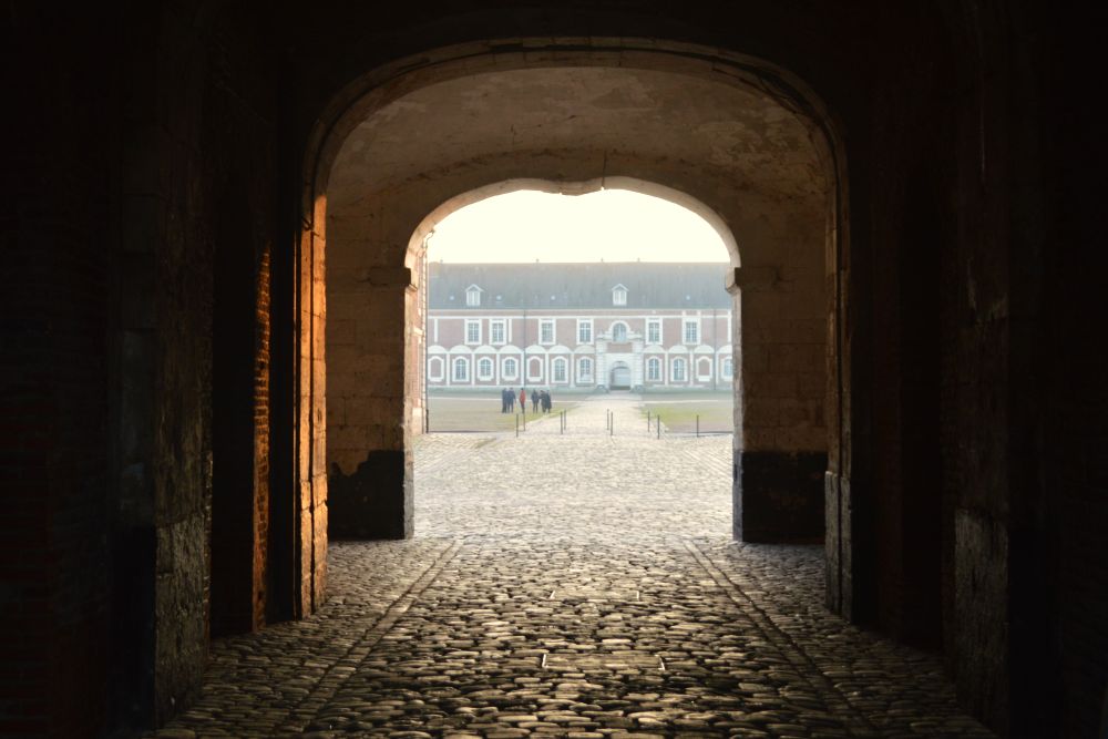 depuis un porche un chemin pavé amène vers sur une place et un bâtiment en briques