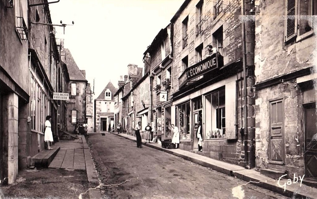 une photographie des années 1940 en noir et blanc d'une rue de village avec des commerces et des habitants