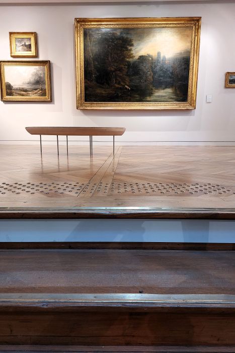 En haut du grand escalier en bois du musée de Valence, un banc attend le visiteur devant des tableaux de paysages