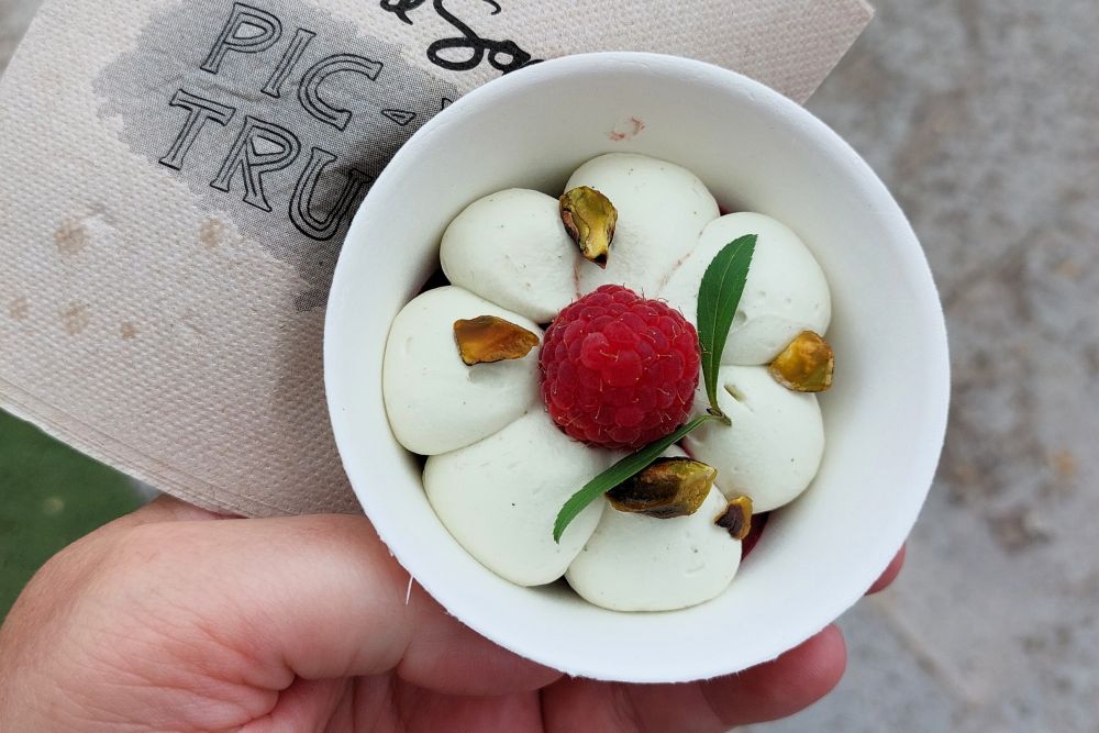 un bol de crème montée surmonté d'une framboise, de pistaches torréfiées et d'une feuille de verveine fraîche, posé sur une serviette en papier du food truck d'Anne Sophie Pic