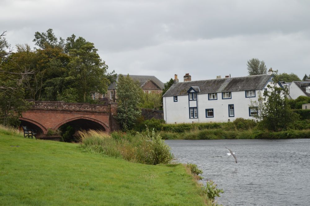 un pont en grès rouge, une maison blanche et une rivière bordée d'une prairie