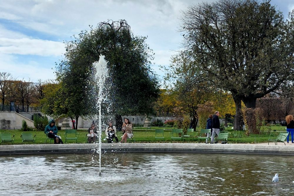 au premier plan, un jet d'eau sur un bassin dans un jardin public, avec en arrière plan des personnes assisses sur les chaises bordant le bassin
